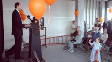 A man stands at a speaking podium, surrounded by orange balloons. He's facing a small crowd of people sitting in folding chairs or standing as they listen to him. A young girl wearing a dress and hair bow waves in the foreground.